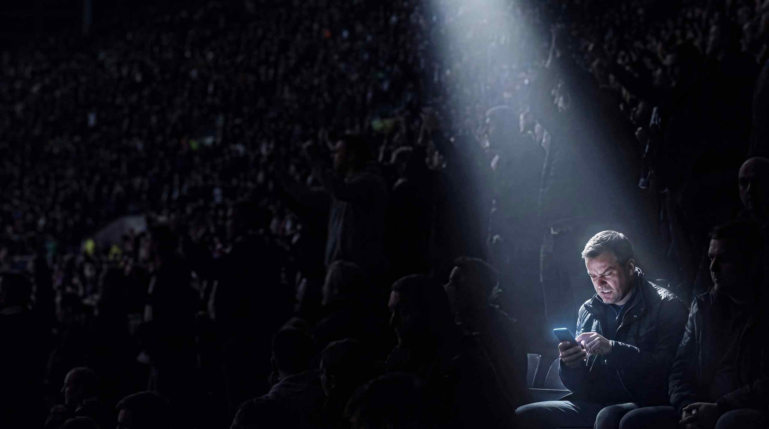 A floodlight on a single man in a stadium looking angrily at his phone.