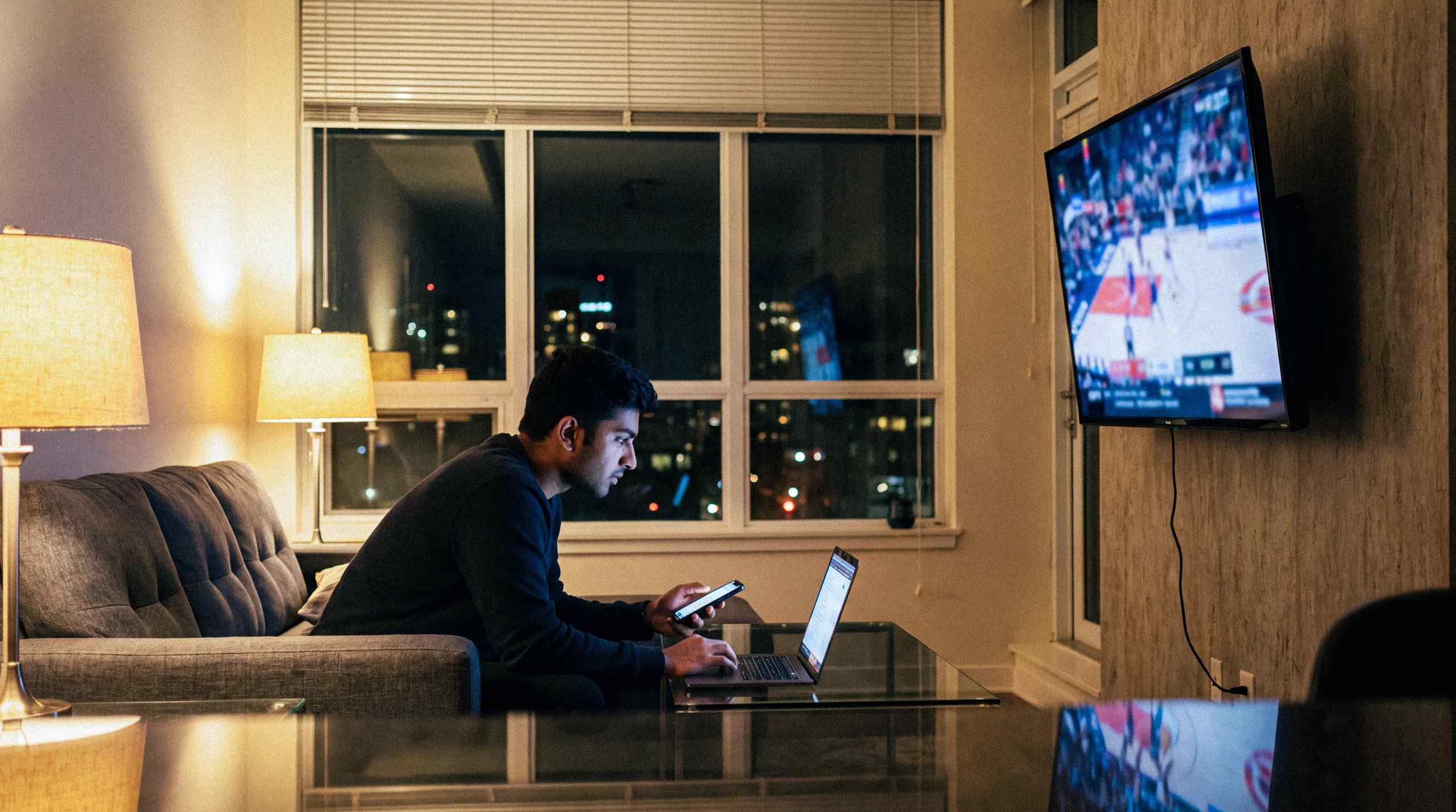 A man watching the NBA at home while placing bets on his mobile phone and laptop.